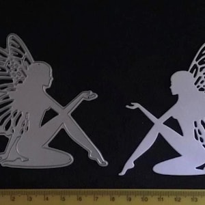 May include: Two silver fairy die cuts. Each fairy is seated with one leg crossed over the other. The fairies have detailed wings and are posed with one arm extended. A ruler with centimetre markings is at the bottom.