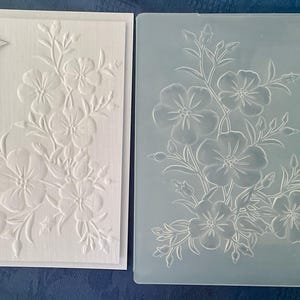 May include: A white square card featuring an embossed floral design and a banner with the text SPECIAL FRIEND, alongside a translucent plastic embossing folder displaying the same floral pattern for creating textured paper crafts.