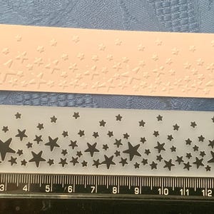 May include: Two rectangular stencils with star patterns. One is a light pink colour with embossed stars. The other is transparent with black star cutouts. A ruler with cm is at the bottom.