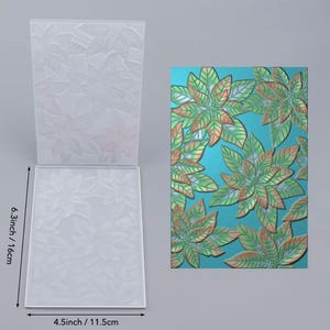 May include: A clear plastic embossing folder featuring a raised poinsettia floral pattern. The folder measures 16 cm by 11.5 cm. Next to it is a sample print showing the embossed design in shades of green and orange on a blue background.