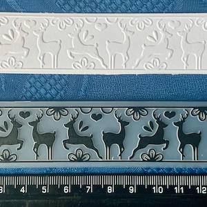 May include: A clear plastic sheet with a raised design of a row of reindeer. The sheet is 15 inches long and has a white background. Below the sheet is a clear plastic stamp with a row of reindeer in a black silhouette. The stamp is 15 centimetres long.