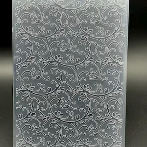 May include: A rectangular, translucent plastic embossing folder featuring a repeating, intricate pattern of swirling vines, delicate leaves, and small buds. The design is recessed into the surface, creating a textured, raised effect for paper crafting projects.