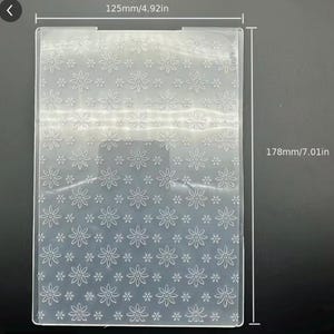 May include: A rectangular clear plastic embossing folder featuring a repeating floral pattern of daisies and small dots. The folder measures 4.92 inches in width and 7.01 inches in height, as indicated by the printed dimension lines and text.
