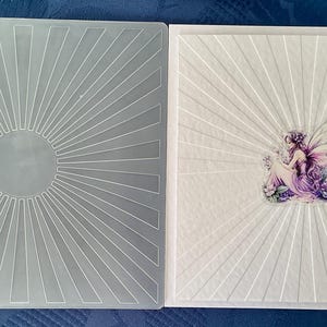 May include: A square translucent plastic embossing folder featuring a sunburst pattern with a central circle. Beside it is a white square cardstock piece embossed with the same sunburst design, featuring a small fairy illustration in the center.
