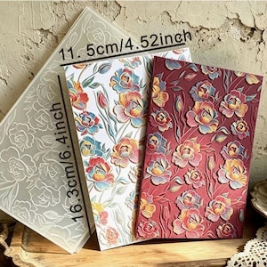 May include: A rectangular embossing folder featuring a raised floral pattern, shown alongside two paper samples with the same design. The folder measures 11.5 cm by 16.3 cm, or 4.52 inches by 6.4 inches. One paper sample is white, and the other is deep red.