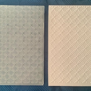 May include: A clear plastic embossing folder with a repeating geometric pattern. The folder is shown next to a piece of paper that has been embossed with the same pattern.