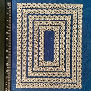 May include: A set of five nested rectangular metal dies with a decorative, woven-style border. The dies are silver-colored and arranged in descending sizes. A ruler with both inches and centimeters is visible along the left side, with the largest die measuring approximately 5.5 inches in length.