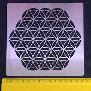 May include: A silver metal stencil featuring a geometric flower of life design. The stencil is square with a flower-like pattern cut out of the metal. A yellow ruler with centimeter markings is at the bottom.