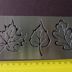 May include: Metal stencil with three leaf designs: a maple leaf, a birch leaf, and an oak leaf. The stencil is approximately 18 cm long.