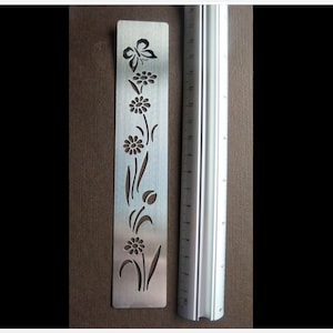 May include: A silver metal bookmark with a floral and butterfly design. The bookmark features cut-out shapes of flowers, leaves, and a butterfly. A metal ruler is next to the bookmark, showing measurements in centimetres.
