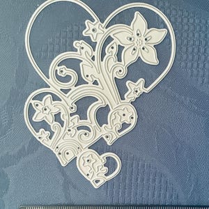 May include: A silver-coloured metal craft die in the shape of a large heart outline filled with intricate floral and star cutouts. A black ruler is positioned at the bottom, showing the die measures approximately 9 centimetres in total width.