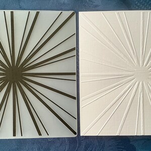 May include: Two square decorative glass tiles. One tile features a white background with a dark brown burst design. The other tile has a white embossed burst pattern. These tiles could be used as coasters or decorative accents.