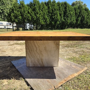 Könnte beinhalten: Ein quadratischer Holztisch mit hellbrauner Tischplatte und beigefarbenem Steinsockel. Der Tisch steht im Freien auf einer Holzplattform, mit einer Rasenfläche und Bäumen im Hintergrund.