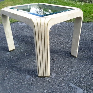 May include: A square, cream-colored side table with a glass top. The table has a unique design with curved corners and brass accents. The legs are rectangular and the table measures approximately 24 inches in height.