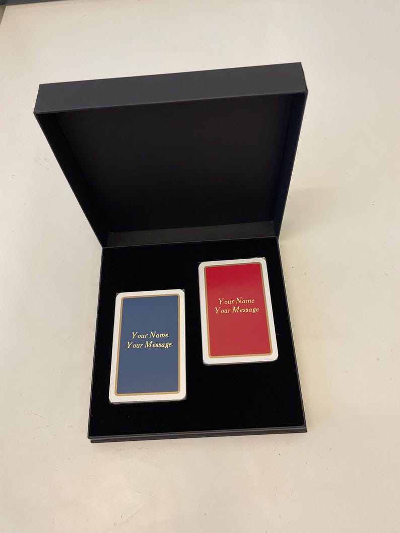 Op de afbeelding: Twee speelkaarten in een zwarte doos. De kaarten zijn blanco en hebben een gouden rand. De bovenste kaart is rood en de onderste kaart is blauw. De tekst op de kaarten luidt "Your Name Your Message".