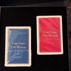 Op de afbeelding: Twee blanco speelkaarten in een zwarte doos. De kaarten zijn wit met gouden randen. E&eacute;n kaart heeft een blauwe achtergrond en de andere een rode. De tekst "Your Name Your Message" is in goud op beide kaarten gedrukt.