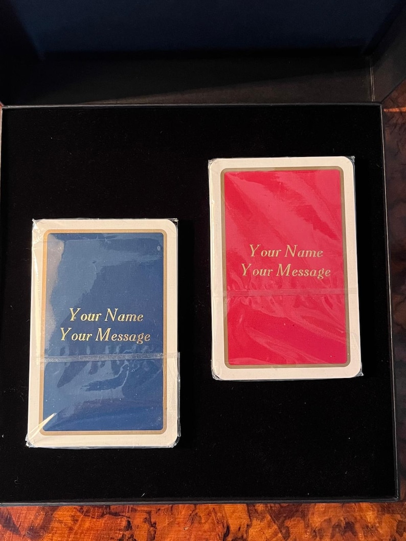 Op de afbeelding: Twee blanco speelkaarten in een zwarte doos. De kaarten zijn wit met gouden randen. E&eacute;n kaart heeft een blauwe achtergrond en de andere een rode. De tekst "Your Name Your Message" is in goud op beide kaarten gedrukt.