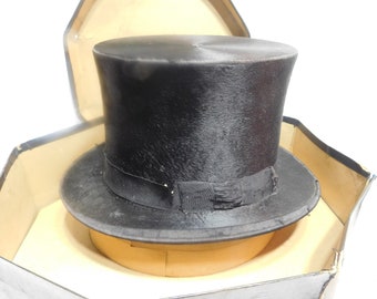 May include: A black top hat with a black ribbon band, presented in a vintage hexagonal hat box. The hat features a tall, cylindrical crown and a wide brim. The box is beige and grey.