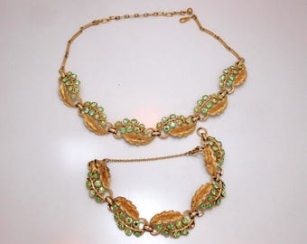 May include: A gold-toned metal necklace and bracelet set featuring a repeating leaf motif. Each leaf link is accented with small, round, light green rhinestones. The necklace includes a thin chain extension with a clasp closure.