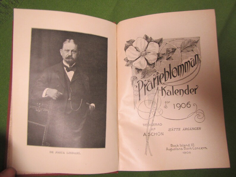 May include: A vintage book cover with a black and white photo of a man in a suit and tie. The cover also features a floral design with the text "Prarieblomman Kalender for 1906" and "Redigerad af A Sch"on Sj"atte Arg"angen".