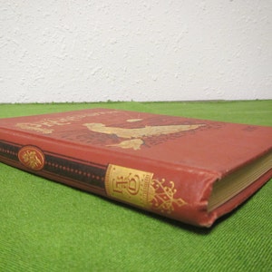 May include: A red hardcover book with gold lettering on the spine and front cover. The book is titled "The Adventures of", and the front cover features an illustration of a woman in a flowing dress.