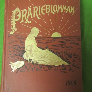 May include: A red book with gold lettering on the cover. The title is "Prarieblomman" and the year 1906 is printed on the bottom right corner. The cover features a woman sitting in a field with a sunset in the background.