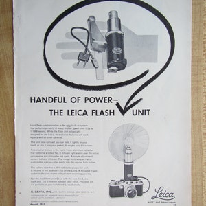 May include: A black and white advertisement for the Leica Flash Unit. The ad features a close-up of the flash unit, which is small and compact. The ad also features a photo of a Leica camera with the flash unit attached. The text on the ad reads "Handful of Power - The Leica Flash Unit".