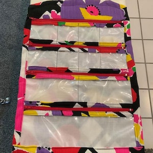 May include: A colorful hanging organizer with multiple clear pockets and red zippers. The fabric features a vibrant pattern of black, pink, yellow, and purple shapes. It has a handle and a pink ribbon for hanging, ideal for storing small items.
