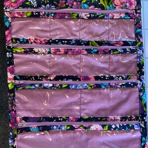 May include: A floral print hanging travel organizer with multiple clear, zippered compartments. The organizer features a dark navy background with pink, purple, and blue flowers. The zippers are purple.