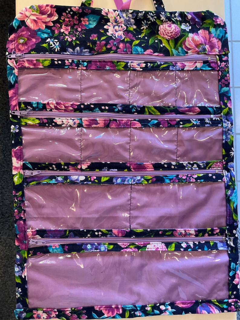May include: A floral print hanging travel organizer with multiple clear, zippered compartments. The organizer features a dark navy background with pink, purple, and blue flowers. The zippers are purple.
