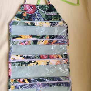 May include: A floral print hanging organizer with multiple clear zippered pockets. The fabric features a pattern of red, yellow, and green flowers on a dark background. It has a handle and a green ribbon for hanging.