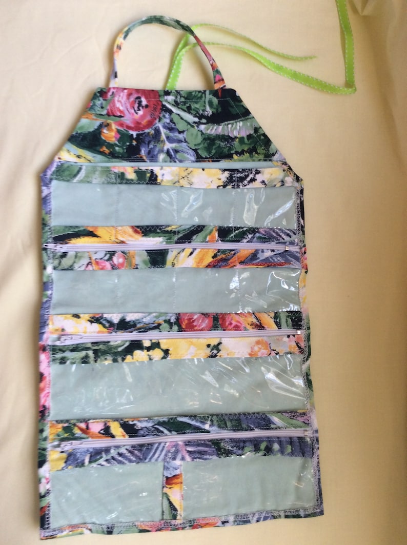 May include: A floral print hanging organizer with multiple clear zippered pockets. The fabric features a pattern of red, yellow, and green flowers on a dark background. It has a handle and a green ribbon for hanging.