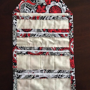 May include: A quilted jewelry travel case with a floral pattern in red, black, and white. The case has multiple zippered compartments and a handle. The tag reads "JEWELRY TRAVEL CASE" and "MADE IN THE USA".