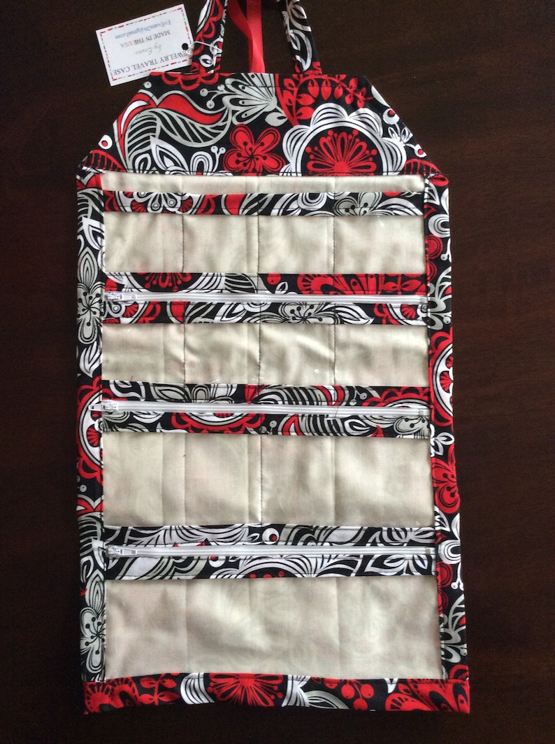 May include: A quilted jewelry travel case with a floral pattern in red, black, and white. The case has multiple zippered compartments and a handle. The tag reads "JEWELRY TRAVEL CASE" and "MADE IN THE USA".