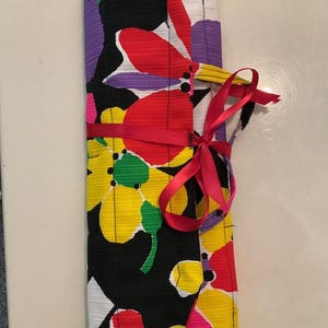 May include: A rolled-up fabric item with a vibrant floral pattern in yellow, red, purple, and white on a black background. A red ribbon is tied around the middle, and a pink ribbon is tied in a bow. The item is likely a case or wrap.