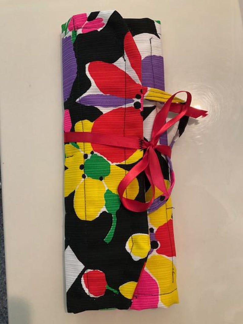 May include: A rolled-up fabric item with a vibrant floral pattern in yellow, red, purple, and white on a black background. A red ribbon is tied around the middle, and a pink ribbon is tied in a bow. The item is likely a case or wrap.