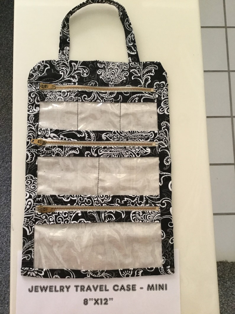 May include: Black and white paisley print jewelry travel case with a handle. The mini case has four zippered compartments with clear plastic windows. The case measures 8" x 12".