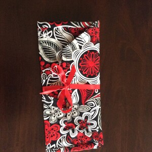 May include: A rectangular fabric item with a floral pattern in red, black, and white, tied with a red ribbon. The design features stylized flowers and leaves. The item is folded and appears to be a case or wrap.