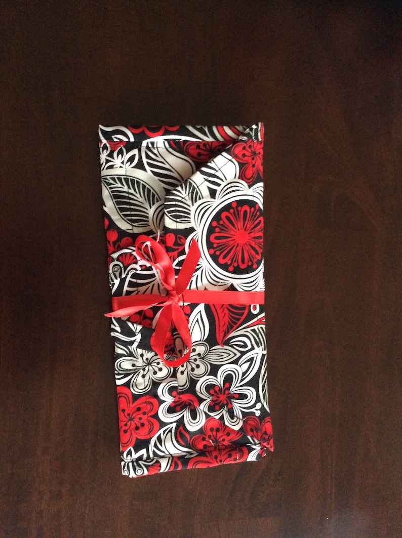 May include: A rectangular fabric item with a floral pattern in red, black, and white, tied with a red ribbon. The design features stylized flowers and leaves. The item is folded and appears to be a case or wrap.
