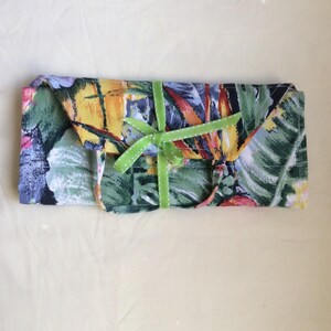 May include: A rectangular fabric item, possibly a case or pouch, with a vibrant floral pattern in green, yellow, red, and blue. A green ribbon with a bow secures the item. The background is a light yellow fabric.