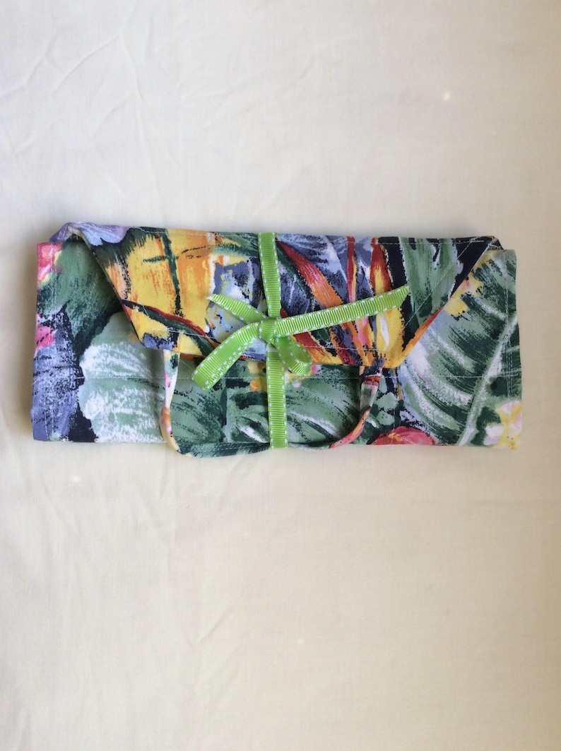 May include: A rectangular fabric item, possibly a case or pouch, with a vibrant floral pattern in green, yellow, red, and blue. A green ribbon with a bow secures the item. The background is a light yellow fabric.