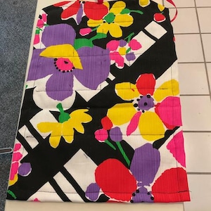 May include: A colorful apron with a floral pattern. The apron features large flowers in shades of purple, yellow, red, and pink against a black and white background. Red ribbon straps are attached at the top.