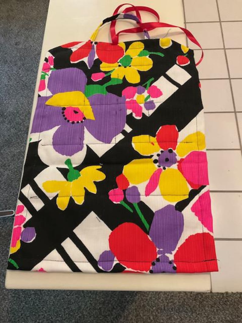 May include: A colorful apron with a floral pattern. The apron features large flowers in shades of purple, yellow, red, and pink against a black and white background. Red ribbon straps are attached at the top.