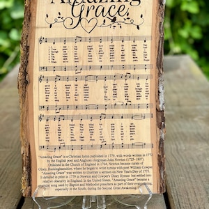 May include: A wooden plaque with the words "Amazing Grace" in elegant script, accompanied by musical notes and a heart design. The plaque displays the hymn's lyrics and history, resting on a clear stand.