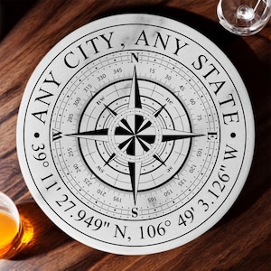 May include: A round, white marble coaster with a compass design. The coaster features the text "ANY CITY, ANY STATE" around the edge, with directional markings and coordinates. The compass rose is black and white.