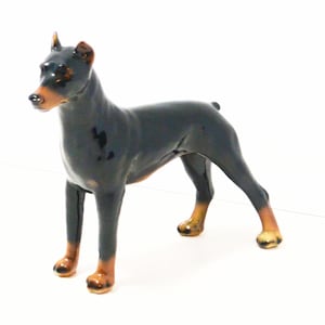 May include: A ceramic figurine of a Doberman Pinscher dog. The dog is black with brown markings on its legs, chest, and face. The figurine is standing in a poised position, with its ears perked up.
