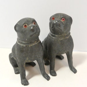 May include: Two black ceramic pug dog figurines with red glass eyes. The dogs are sitting with their heads tilted up.