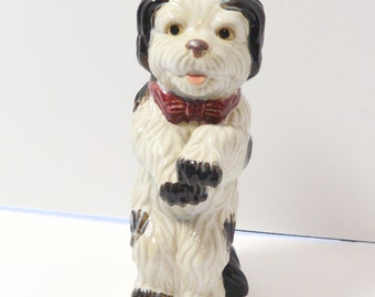 May include: A ceramic figurine of a black and white dog wearing a red bow tie. The dog is standing on its hind legs with its front paws raised.