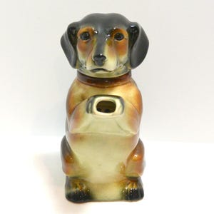 May include: A ceramic dachshund figurine, featuring a brown and black color scheme. The dog is in a seated position with a small opening in its chest. The figurine has a glossy finish and is likely a decorative item.