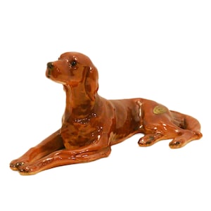 May include: A glossy, reddish-brown ceramic figurine of a dog in a lying position. The dog has long, floppy ears, a dark nose, and dark eyes. A small, gold-colored oval sticker is visible on the dog's back, near the hindquarters.